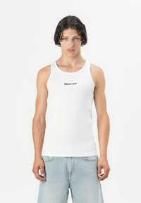 Hvit ribbet singlet med rund hals og tynne skulderstropper, prydet med subtil svart tekst brodert på brystet. Brukt sammen med lyseblå jeans.