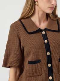 Mulher vestindo uma blusa curta texturizada castanha com bordas pretas, botões dourados e bolsos frontais, mostrando o perfil lateral do torso superior e da cabeça.