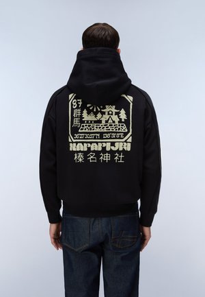 Mann trägt schwarzen Hoodie mit beiger Grafik von Häusern, Bäumen, Koordinaten und japanischem Text auf dem Rücken, kombiniert mit dunkelblauen Jeans.