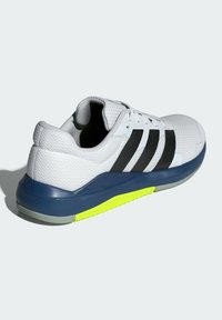 Zapatilla deportiva blanca con parte superior de malla texturizada, rayas negras y suela de un azul oscuro. Acentos amarillos brillantes en la suela ofrecen contraste.