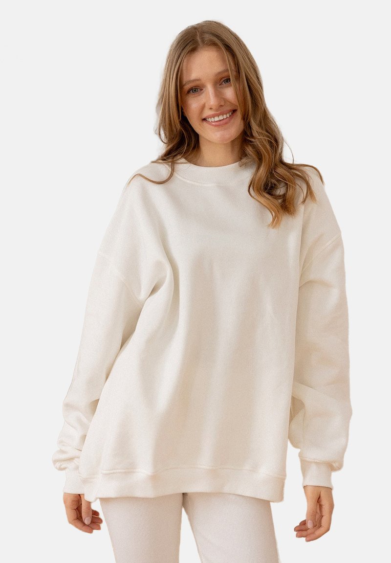 Przesadnie duża, długorękawowa bluza w kolorze off-white. Posiada ściągacz przy dekolcie i mankietach, gładką fakturę i brak widocznych wzorów.