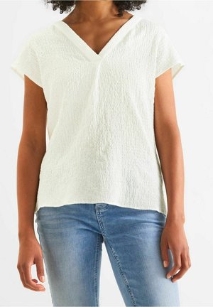 Vrouw die een wit textuur kortermig V-hals shirt draagt, gecombineerd met lichtblauwe jeans, staand tegen een effen witte achtergrond.