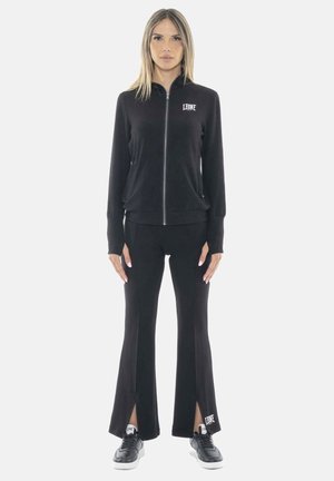 Femme debout portant une veste noire zippée et un pantalon évasé avec des fentes, ainsi que des baskets noires, tous portant la marque « Leone ».