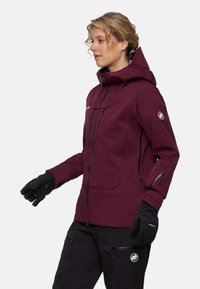 Vinröd vattentät jacka med huva, framdragkedja, sidofickor och justerbara ärmslut, kombinerad med svarta handskar. Mjuk textur och figursydd design.