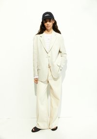 Blazer in lino color beige chiaro, camicia a maniche lunghe a righe, pantaloni ampi color crema, sandali neri e berretto blu navy con testo bianco.
