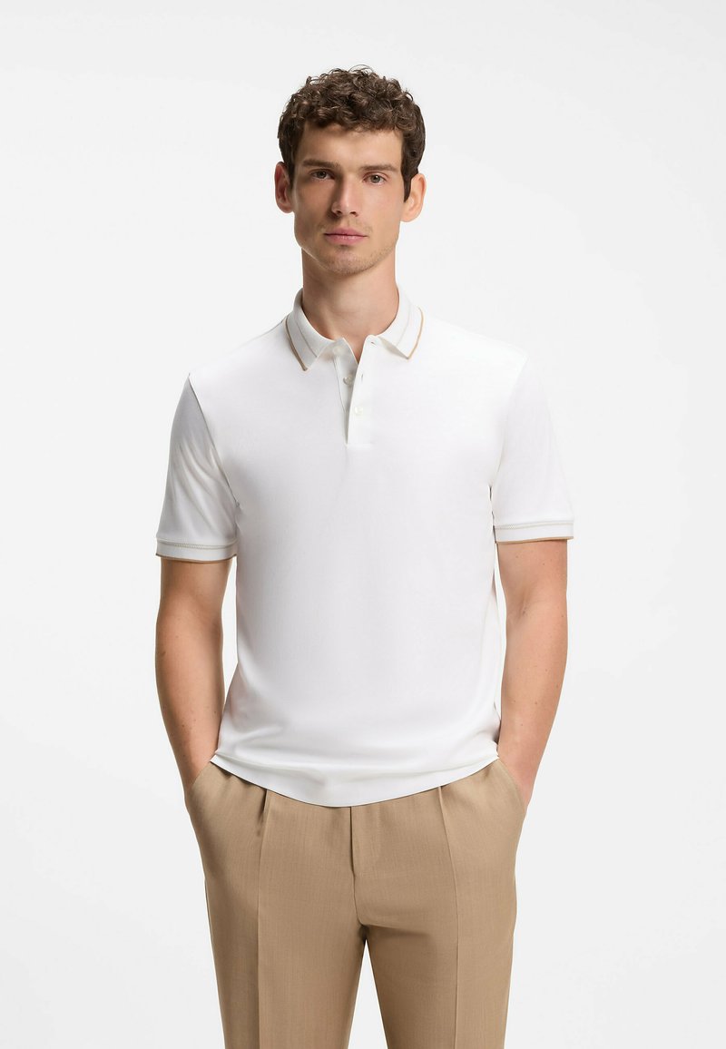 Jeune homme aux cheveux bouclés bruns, portant un polo blanc à manches courtes et un pantalon beige, mains dans les poches, fond neutre.