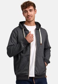 Sweat à zip gris foncé avec une texture, cordons blancs, poches latérales et poignets côtelés. Porté par-dessus un T-shirt blanc.