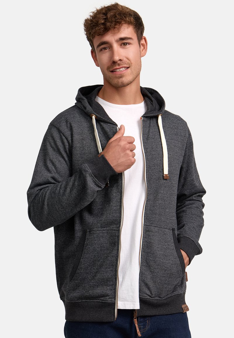 Dunkelgrauer Zip-Hoodie mit strukturiertem Aussehen, weißen Kordeln, seitlichen Taschen und gerippten Bündchen. Über einem weißen T-Shirt getragen.