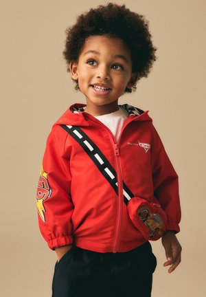 Young child with curly hair wearing a red Cars-themed jacket with zipper and a matching Cars-shaped crossbody bag, smiling and looking to the side.