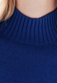 Blauwe gebreide trui met een geribbelde hoge hals. De textuur is glad met strakke stiksels, en toont een solide, levendige kleur.