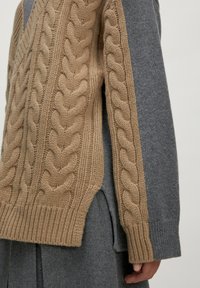 Close-up van een persoon die een beige chunky kabelgebreide trui draagt, gelaagd over een grijze wollen langemouwen kledingstuk met een ribbelmanchet.