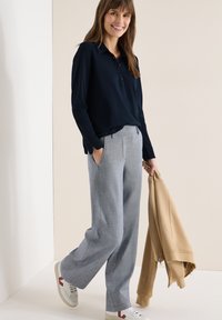 Femme souriante marchant à l'intérieur, portant un haut bleu marine à manches longues, un pantalon gris à jambes larges, des baskets blanches, tenant une veste zippée beige dans une main.