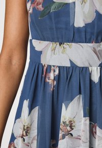 Květinové šaty s modrou látkou s velkými bílými a růžovými květinovými vzory, s úzkým pasem a nařasenou sukní pro větší objem.