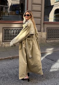 Khaki trench coat with a belted waist, wide sleeves, and a smooth, shiny texture. Worn over black high-heeled sandals on a city street.