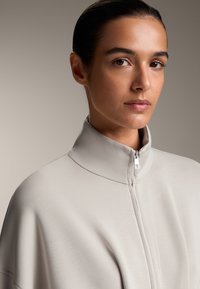 Vaaleanharmaa korkean kauluksen takki, joka on valmistettu sileästä kankaasta. Siinä on edessä vetoketju ja rento istuvuus. Pehmeä pinta ja minimalistiset muotoiluyksityiskohdat.