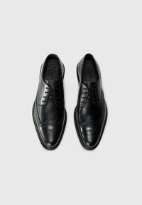 Chaussures habillées en cuir noir avec un design brogue, bout pointu et finition brillante. Comprend un système de laçage et des perforations décoratives.