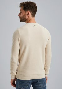 Sweatshirt texturé crème avec un col rond, des poignets et un ourlet côtelés, présentant un petit détail de bouton au dos. Porté avec un jean bleu.