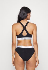 Svart trenings-BH og high-waisted truser med kryss-bak design. BH-en har en hvit kant med "GUESS" tekst. Glatt stofftekstur.