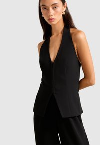 Woman with long dark hair wearing gold hoop earrings and a sleeveless black V-neck halter top with matching black pants.