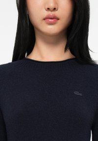 Mladý človek so rovnými čiernymi vlasmi, ktorý má na sebe námornícky modrý sveter s malým vyšitým logom krokodíla na hrudi.