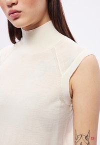 Krém színű, ujjatlan, állónyakú felső könnyű, enyhén áttetsző anyagból. Látható varrásrészletek a vállakon és a nyakkivágásnál.