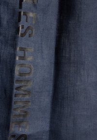 Denimstof in een diepe blauwe kleur met een verticale tekstpatroon dat "HOMMES" leest. De textuur lijkt glad met een subtiele glans.