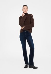 Maglione a collo alto a coste marrone con un motivo a rombi, dotato di bottoni decorativi sulle spalle, abbinato a jeans a zampa d'elefante blu scuro e stivali neri.