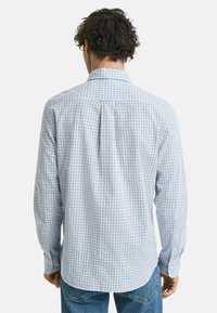 Mann mit lockigem Haar, der ein langärmliges, weißes Hemd mit blauem Karomuster und blaue Jeans trägt, von hinten gezeigt.
