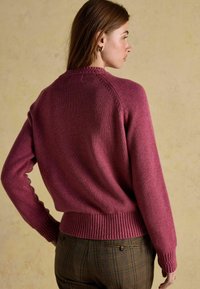 Naine, kellel on seljas keha rõhutav lilla kudum ja pruun ruuduline pükskostüüm, on nähtav tagantpoolt, seistes beežil taustal.