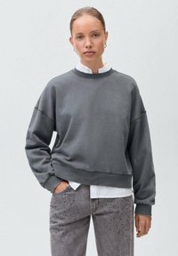 Siva zipka s krojenjem, širokimi rokavi in okroglim izrezom, v kombinaciji z svetlo sivo jeans hlačami z diskretnimi okraski.