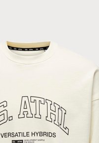 Creme sweatshirt med rund halsudskæring, der har fed sort tekst. Stoffet ser glat og blødt ud, med detaljer i trimmet indeni kraven.