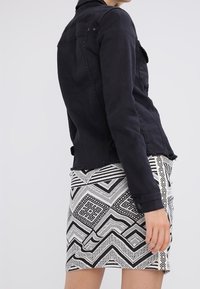Giacca di denim nera con dettagli in bottone, design corto e orlo sfrangiato; indossata sopra una gonna geometrica nera e bianca a motivi.