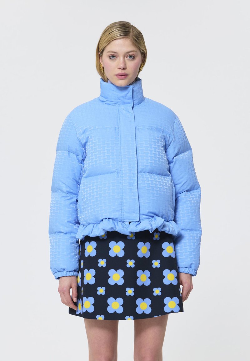 Jachetă puffer bleu deschis cu model texturat și tiv ondulat, purtată cu o fustă neagră cu imprimeu floral albastru și galben.
