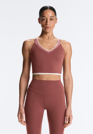 Frau steht, trägt ein passendes mauvefarbenes Sport-BH und hoch taillierte Leggings, mit neutralem Ausdruck vor einem einfarbigen Hintergrund.