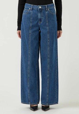 Person iført højtaljede vide blå jeans med lodrette syninger og sorte spidse sko mod en ensfarvet baggrund.