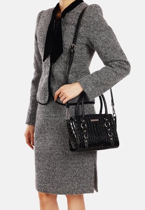 Borsa nera in pelle sintetica con texture a coccodrillo, dettagli in metallo argentato e doppio manico superiore, tenuta accanto a un completo in tweed grigio.