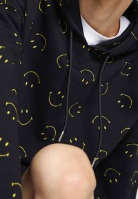 Black hoodie featuring a pattern of yellow smiley faces, with drawstrings and a soft texture. Visible is a person's knee.