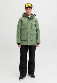 Grüne isolierte Jacke mit mehreren Fronttaschen, schwarzen Akzenten und Kapuze, kombiniert mit schwarzen Hosen und einer Skibrille. Glatte Textur.