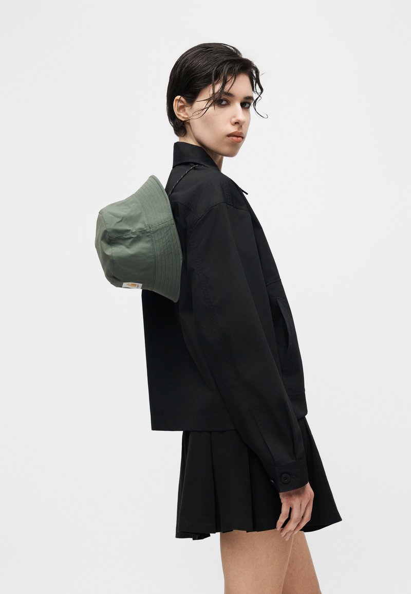 Giovane con capelli corti bagnati che indossa una giacca nera e una gonna, con un cappello a secchiello verde appeso sulla schiena.