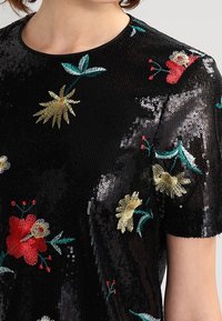 Top noir à sequins avec broderie florale colorée en rouge, or et vert. Manches courtes et col rond. Surface texturée.