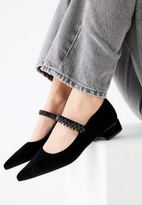 Zapatos Mary Jane de terciopelo negro con punta afilada, adornados con una correa de cuentas alrededor del tobillo y un pequeño tacón cuadrado.