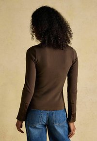 Brown ribbed long-sleeve top with fitted design, buttoned cuffs, and a round neckline, paired with high-waisted blue jeans.