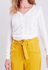 Bílý pletený kardigan s výstřihem do V, zdobený vzorem listů a zapínáním na knoflíky, zkombinovaný s vysokými žlutými kalhotami s opaskem.