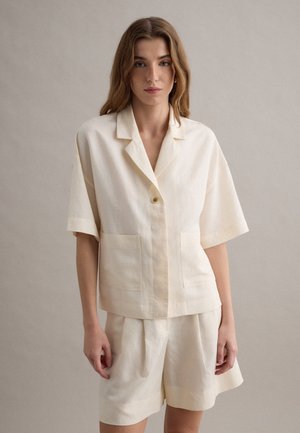 Giovane donna con capelli lunghi e mossi indossa una camicia crema a maniche corte con bottoni e pantaloncini larghi coordinati, in piedi davanti a uno sfondo semplice.