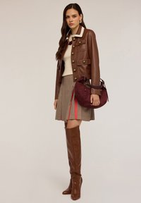Donna che indossa una giacca di pelle marrone, un top crema, una gonna a quadri con strisce rosse, stivali alti fino al ginocchio marroni, mentre tiene una borsa di camoscio bordeaux.