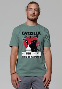 T-shirt vert à manches courtes avec un graphique d'un chat noir contre un cercle rouge, avec le texte "CATZILLA" et "KING OF PAWSTER" au-dessus et en dessous.