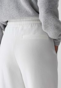 Weiße Sweatpants mit elastischem Bund und einer flachen Gesäßtasche. Der Stoff wirkt weich mit einer glatten Textur. Ein grauer Strickpullover ist teilweise sichtbar.