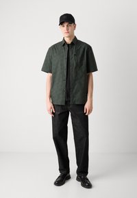 Korte mouwen, donkergroene gemusterde shirt, zwarte t-shirt eronder, gecombineerd met donkere spijkerbroek en zwarte schoenen, aangevuld met een zwarte pet.