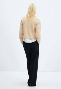 Gekürzter beiger Pullover mit gerippten Bündchen, über einem weißen Hemd getragen, kombiniert mit hoch taillierten schwarzen Hosen. Glatte Textur, minimalistisches Design.