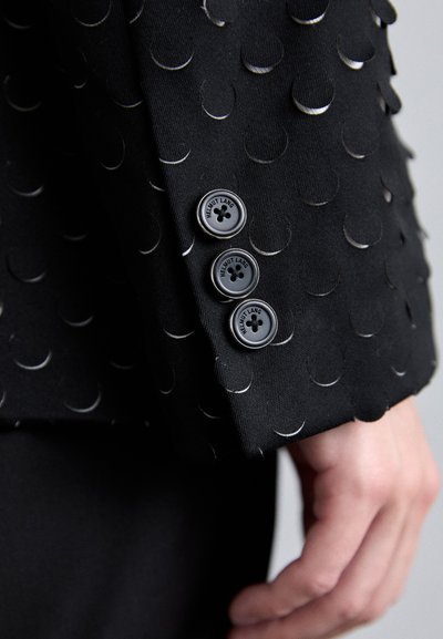 Black jacket sleeve featuring textured fabric adorned with circular silver accents. Three black buttons with "Helmut Lang" insignia at cuff.